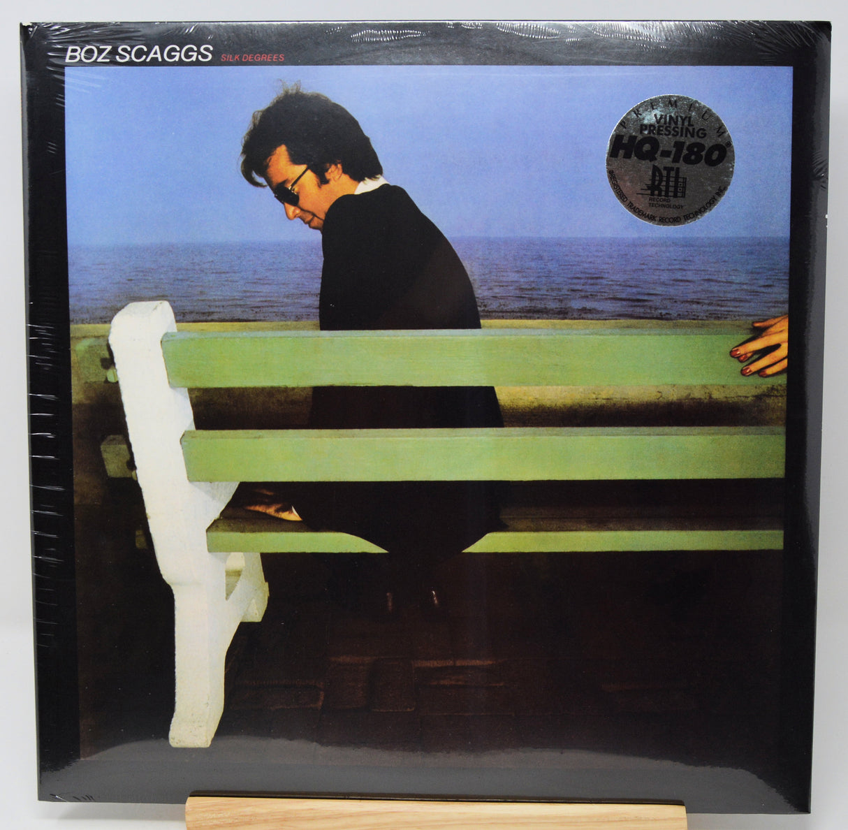 Boz Scaggs '66 Memories' LP record album in shrink wrap, displayed on a wooden stand. The cover art features Boz Scaggs sitting on a green bench by the sea.