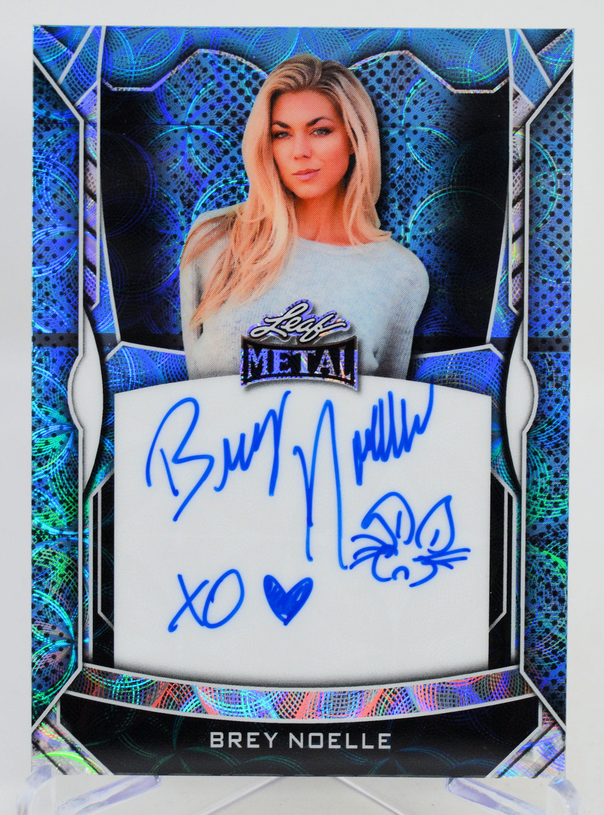 Autographed card of a person wearing a shirt with 'Metal' branding, set against a blue and black decorative border.