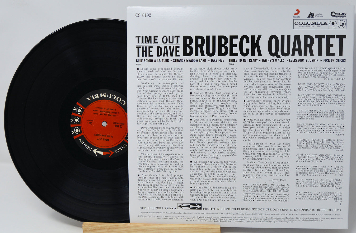 A vinyl record partially pulled from its sleeve, showing the black record with a red-and-black Columbia center label beside the printed album cover for Time Out: The Dave Brubeck Quartet. Visible brands include Columbia and the Brubeck Quartet.