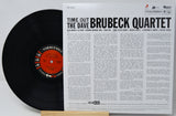 A vinyl record partially pulled from its sleeve, showing the black record with a red-and-black Columbia center label beside the printed album cover for Time Out: The Dave Brubeck Quartet. Visible brands include Columbia and the Brubeck Quartet.