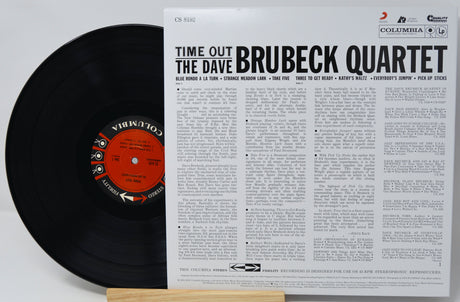 A vinyl record partially pulled from its sleeve, showing the black record with a red-and-black Columbia center label beside the printed album cover for Time Out: The Dave Brubeck Quartet. Visible brands include Columbia and the Brubeck Quartet.