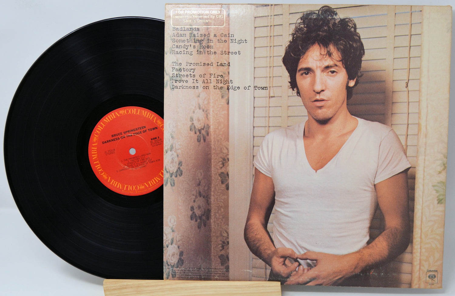 A vinyl record partially pulled from its sleeve, showing the black record with a bright orange center label and the printed album cover featuring a man in a white V-neck shirt against beige shutters.