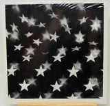 A square black art print or canvas featuring a scattered pattern of white stars, shown wrapped in protective plastic with a wooden display block visible at the bottom.