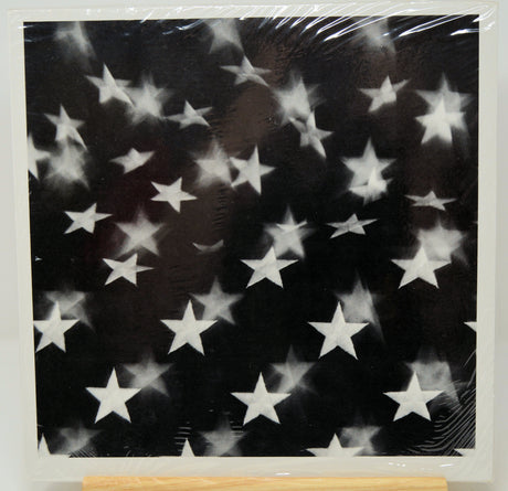 A square black art print or canvas featuring a scattered pattern of white stars, shown wrapped in protective plastic with a wooden display block visible at the bottom.