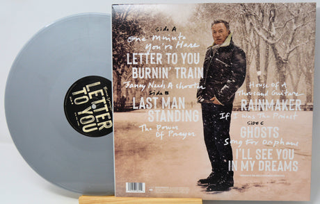 A vinyl record partially visible behind its album cover, photographed on a light wood surface. The record is pale translucent gray, and the cover art shows a man standing outdoors in a snowy scene with white handwritten-style track titles over a sepia-toned background.