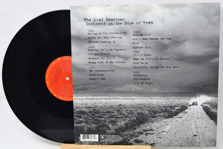 Vinyl record album shown with the black LP partially pulled from the back cover, which features a black-and-white landscape photo and the title 'The Lost Sessions: Darkness on the Edge of Town.'.