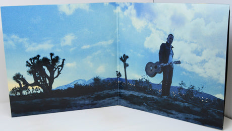 A large printed panel or backdrop featuring a desert landscape scene with a person holding a guitar under a blue cloudy sky, photographed against a white studio background.