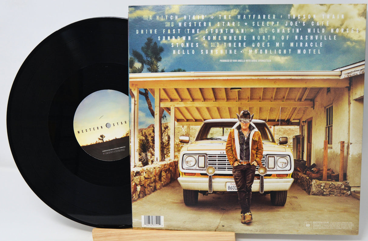 A vinyl record partially pulled from its sleeve, showing the black disc on the left and the album cover on the right. The cover features a man standing in front of a white car under a blue sky, with the text 'Hitch Hike' and tracklist visible at the top.