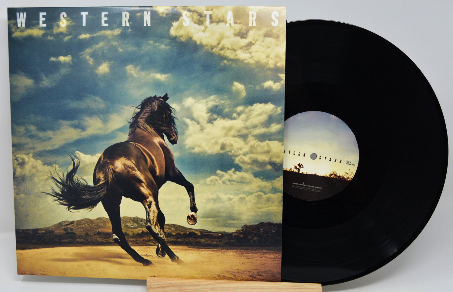 A vinyl record partially pulled from its sleeve, showing the album cover art of a rearing horse under a dramatic cloudy sky. The black record is visible on the right, with the Western Stars title printed on both the cover and the center label.