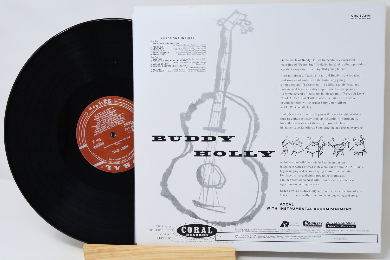 A vinyl record partially pulled from its sleeve, showing the black record with a red-orange label next to the white printed inner sleeve featuring a guitar illustration and the text 'BUDDY HOLLY' and 'Coral Records'.