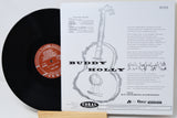 A vinyl record partially pulled from its sleeve, showing the black record with a red-orange label next to the white printed inner sleeve featuring a guitar illustration and the text 'BUDDY HOLLY' and 'Coral Records'.