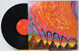 A vinyl record partially tucked behind its illustrated album cover, photographed on a plain light background. The cover artwork is a vivid psychedelic scene with orange, pink, yellow, purple, and brown tones, and the record itself is black with a bright orange center label.