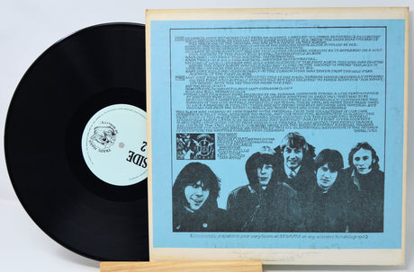 A vinyl record partially pulled from its wooden sleeve, showing the black record and the back cover insert with a light blue background, text, and a black-and-white band photo.