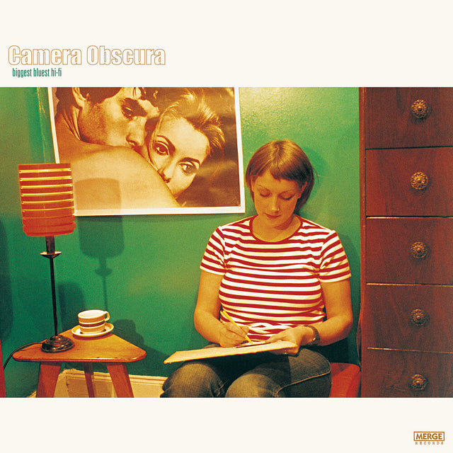 Album cover with a person sitting on a stool, writing, with a lamp and poster in the background.