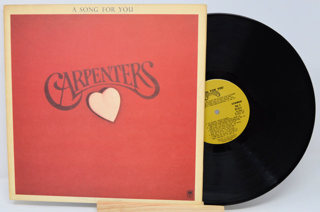 Vinyl record with red cover featuring 'Carpenters' and a heart, on a light gray background.