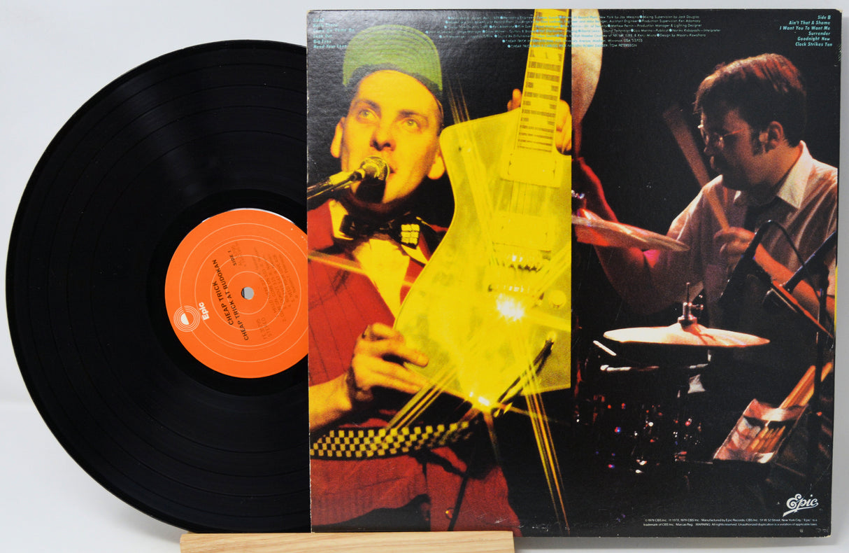 A vinyl record partially pulled from its album cover, showing a black record with an orange center label and a printed cover featuring a guitarist and drummer against a dark background.