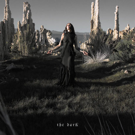 A woman in a long black dress stands in a grassy, rocky outdoor landscape with tall tufa-like rock formations behind her, arms outstretched. The scene is desaturated and moody, with the text 'the dark' at the bottom.