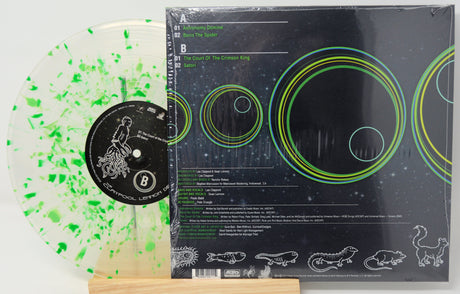 A vinyl record album shown with the back cover and part of the translucent record. The record is clear with bright green paint splatter, and the cover is black with green and yellow circular graphics and track listing text.