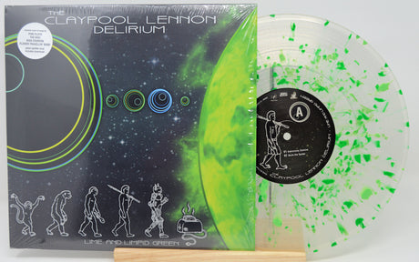 The Claypool Lennon 'Delirium' vinyl record album with its illustrated cover jacket and a translucent green-and-white record partially visible behind it, displayed on a small wooden stand.