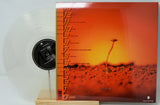 Vinyl record album cover and a translucent clear vinyl record displayed on a small wooden stand. The cover features a vivid orange and red desert landscape with a small plant silhouette and vertical black text along the left edge.