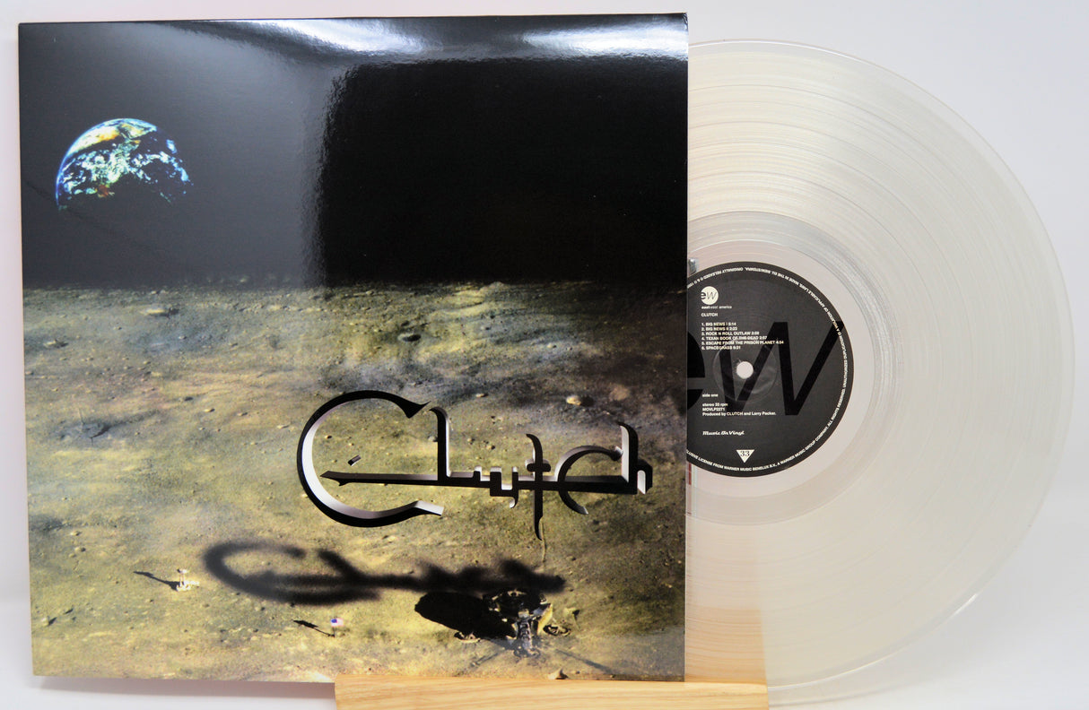 A vinyl record album with its cover jacket standing beside the translucent clear record on a small wooden stand. The cover artwork shows a dark space-like scene with a small Earth in the upper left and stylized metallic lettering across the center.