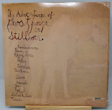 A vinyl record album cover for 'The Adventures of Ghosthorse and Stillborn' on a cardboard display stand. The cover is a warm tan/beige color with handwritten black ink text listing various animal names, and the record sleeve is wrapped in protective plastic.