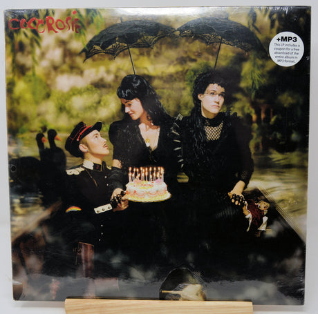 Cocteau Blues vinyl record LP cover on a wooden display stand, featuring three women in black Victorian-style clothing under black lace umbrellas in a swampy outdoor scene, with a birthday cake and an MP3 sticker overlay.