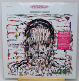 Vinyl record album cover for 'coltrane's sound' with a white background, abstract multicolor portrait artwork, a pink sticker on the right, and a small wooden display stand visible at the bottom.