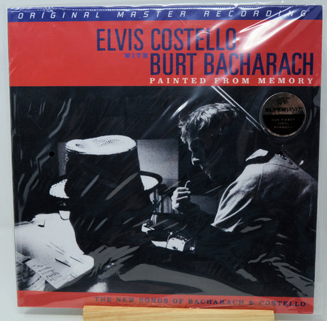 Elvis Costello and Burt Bacharach 'Painted from Memory' vinyl record album cover, sealed in plastic shrink wrap, displayed on a wooden stand against a white background.