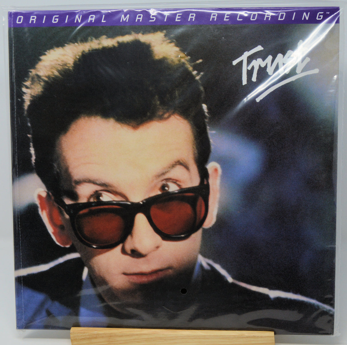 A vinyl record sleeve for Trust's 'Original Master Recording' album, featuring a close-up portrait of the artist wearing dark sunglasses, displayed on a small wooden stand.