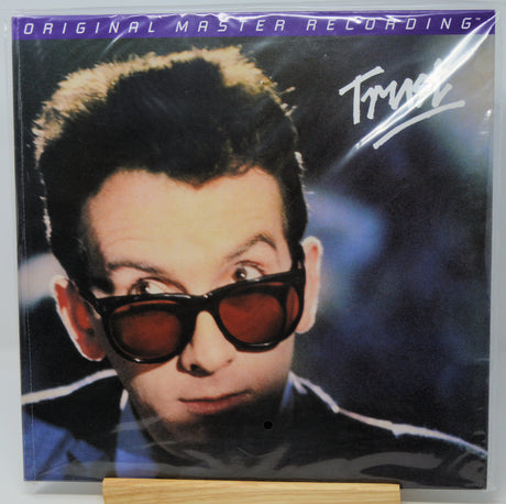 A vinyl record sleeve for Trust's 'Original Master Recording' album, featuring a close-up portrait of the artist wearing dark sunglasses, displayed on a small wooden stand.