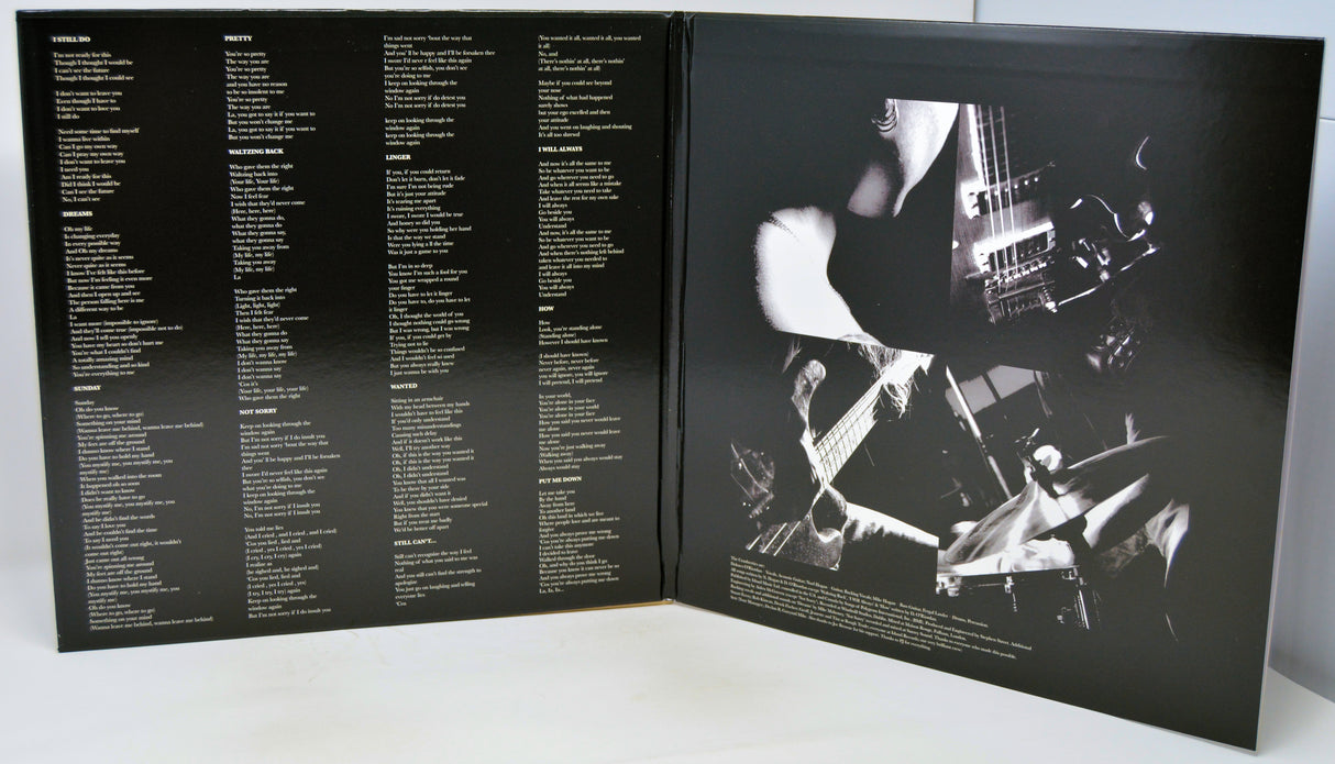 An open black booklet or album insert photographed from above on a white surface. The left page contains multiple columns of small white text, and the right page shows a high-contrast black-and-white photo of a person playing a guitar with drum sets in the background.