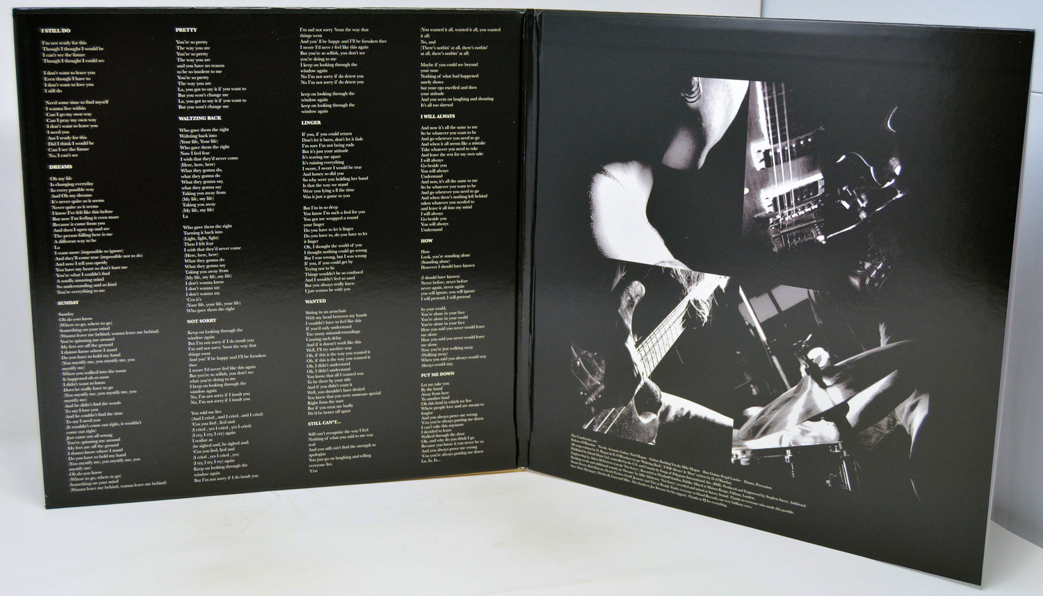 An open black booklet or album insert photographed from above on a white surface. The left page contains multiple columns of small white text, and the right page shows a high-contrast black-and-white photo of a person playing a guitar with drum sets in the background.