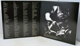 An open black booklet or album insert photographed from above on a white surface. The left page contains multiple columns of small white text, and the right page shows a high-contrast black-and-white photo of a person playing a guitar with drum sets in the background.