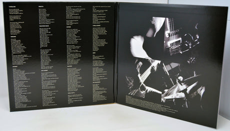 An open black booklet or album insert photographed from above on a white surface. The left page contains multiple columns of small white text, and the right page shows a high-contrast black-and-white photo of a person playing a guitar with drum sets in the background.