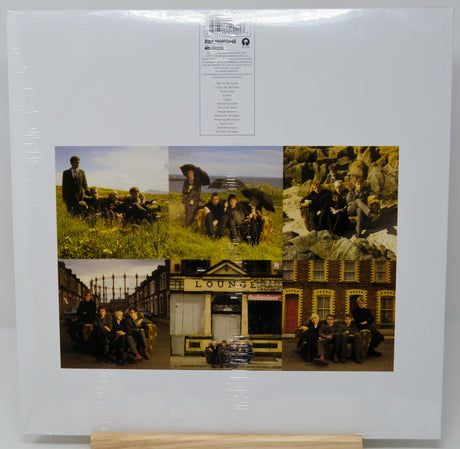A square album cover for 'Best of Ireland 2000' by the band U2, still sealed in its original shrink wrap and displayed on a small wooden stand. The cover features a collage of six color photographs depicting the band members in various outdoor and urban settings, with a white background and a white tracklist label at the top.