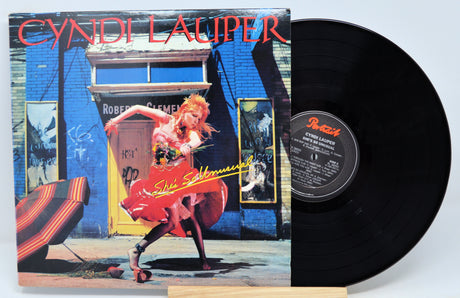 Cyndi Lauper - She's So Unusual vinyl record album cover featuring Cyndi Lauper in a red dress sitting on a bench with a cityscape in the background.