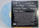 A vinyl record package showing the back of a black album sleeve wrapped in plastic, with a light blue marbled vinyl record partially visible behind it on a white background.