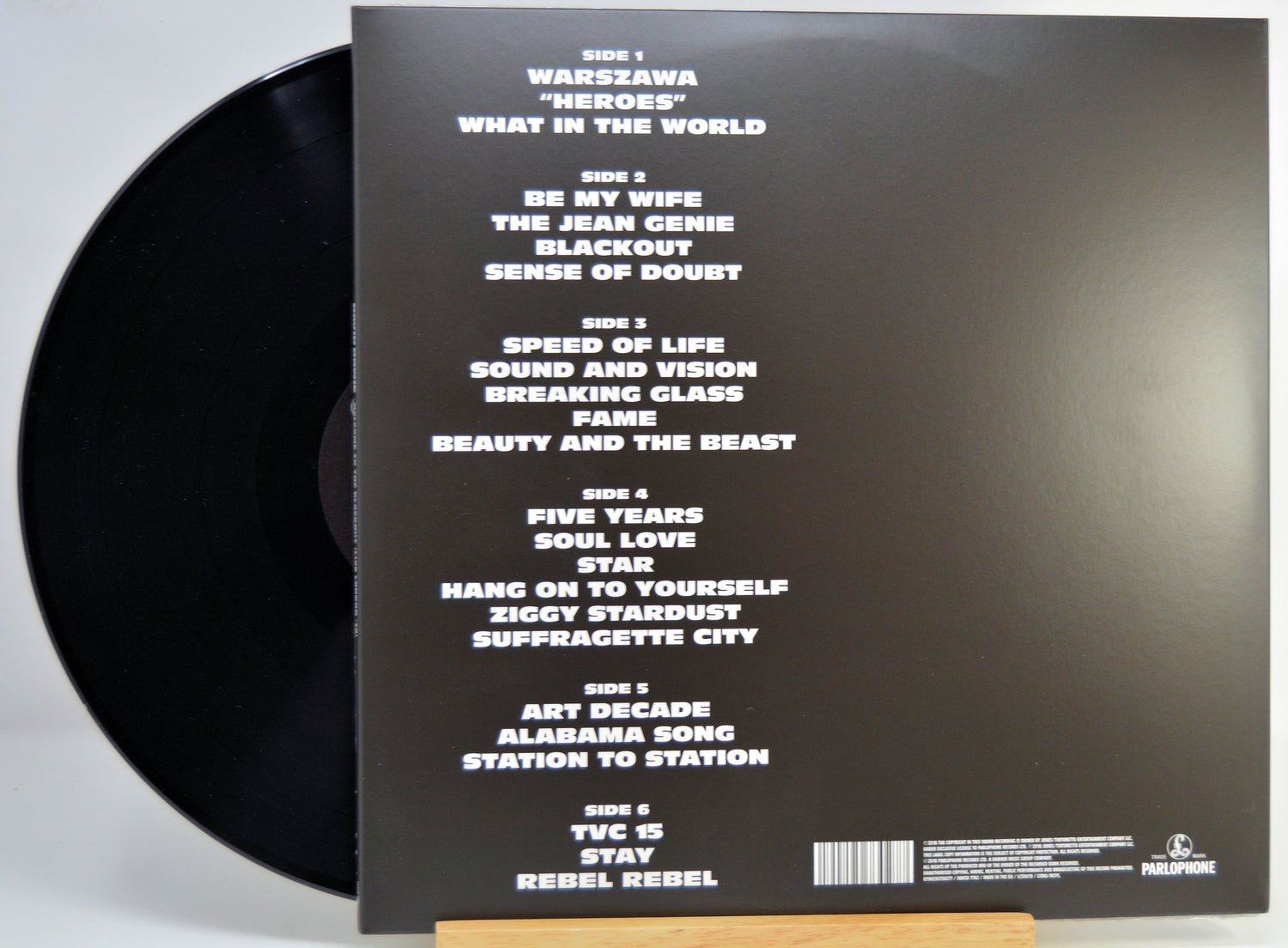 The back of a vinyl record sleeve displayed on a small wooden stand, showing the tracklist in white text on a black background with part of the black record visible at the left edge.