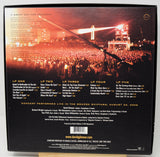 The back cover of a David Gilmour live album print, showcasing a live concert performance in the Grabski Shipyard on August 29, 2006. The design features a dark background with a vibrant orange sunset sky, a crowd of concertgoers, and a bridge in the distance, with track listings and credits printed in white and gold text.