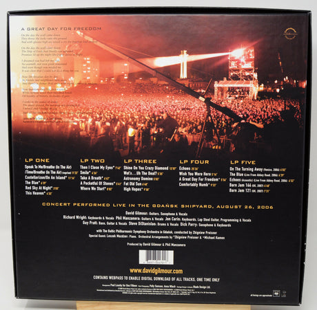 The back cover of a David Gilmour live album print, showcasing a live concert performance in the Grabski Shipyard on August 29, 2006. The design features a dark background with a vibrant orange sunset sky, a crowd of concertgoers, and a bridge in the distance, with track listings and credits printed in white and gold text.