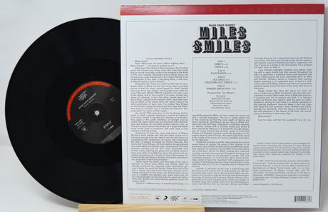 A vinyl record partially pulled from its sleeve, showing the back of the packaging with track listing and text. The record is black with a red and black center label, and the sleeve is white with a red top border and black 'Miles Smiles' title.