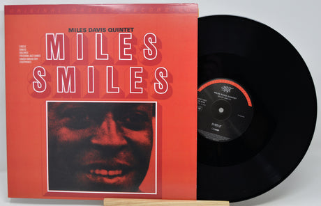 Miles Davis Quintet 'Miles Smiles' vinyl record album with the orange-red cover jacket standing next to a black LP record on a light gray background.