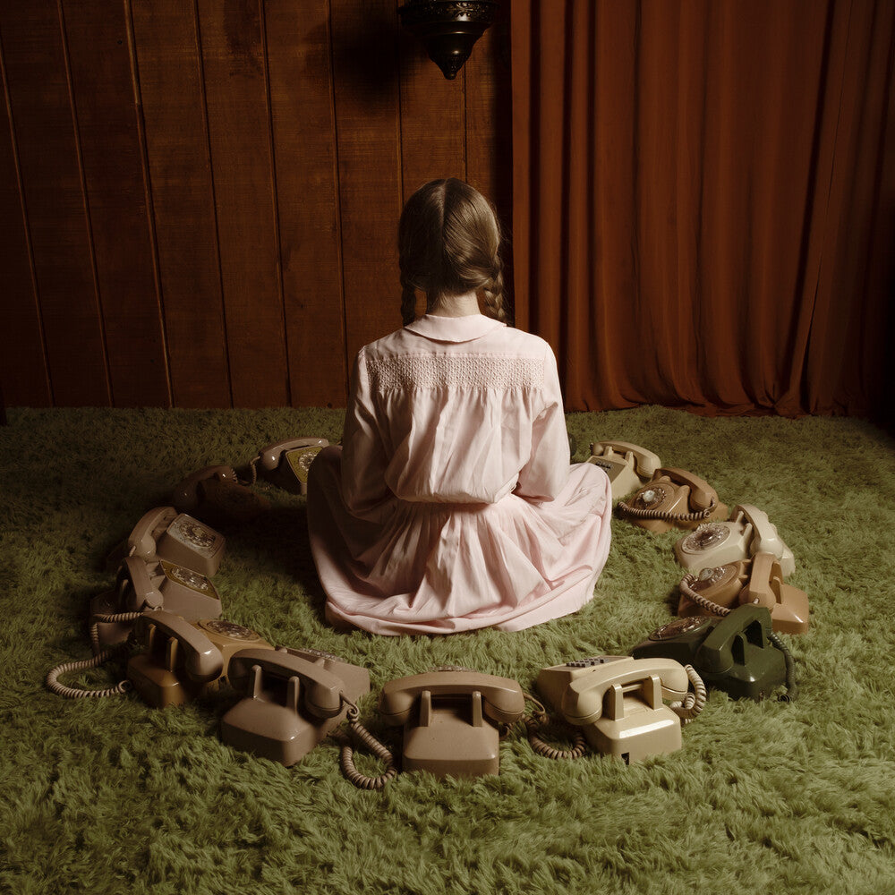 A young girl with braided hair sits on a green shag rug, viewed from behind, wearing a light pink long-sleeve dress. She is surrounded by a circle of vintage rotary phones against a dark wood-paneled wall and brown curtains.