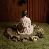 A young girl with braided hair sits on a green shag rug, viewed from behind, wearing a light pink long-sleeve dress. She is surrounded by a circle of vintage rotary phones against a dark wood-paneled wall and brown curtains.