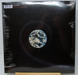 A vinyl record album cover on a black background, wrapped in clear plastic shrink wrap and displayed upright on a small wooden stand. The center features a circular Earth-like globe graphic in blue, white, and gray tones.