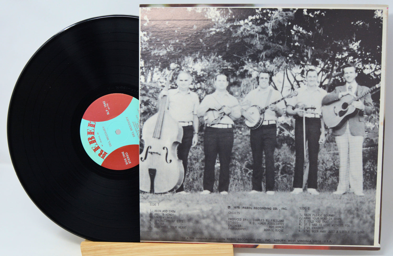 A vinyl record partially pulled from its sleeve, showing a black LP with a red and turquoise center label next to a black-and-white album cover featuring five musicians standing outdoors.