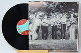 A vinyl record partially pulled from its sleeve, showing a black LP with a red and turquoise center label next to a black-and-white album cover featuring five musicians standing outdoors.