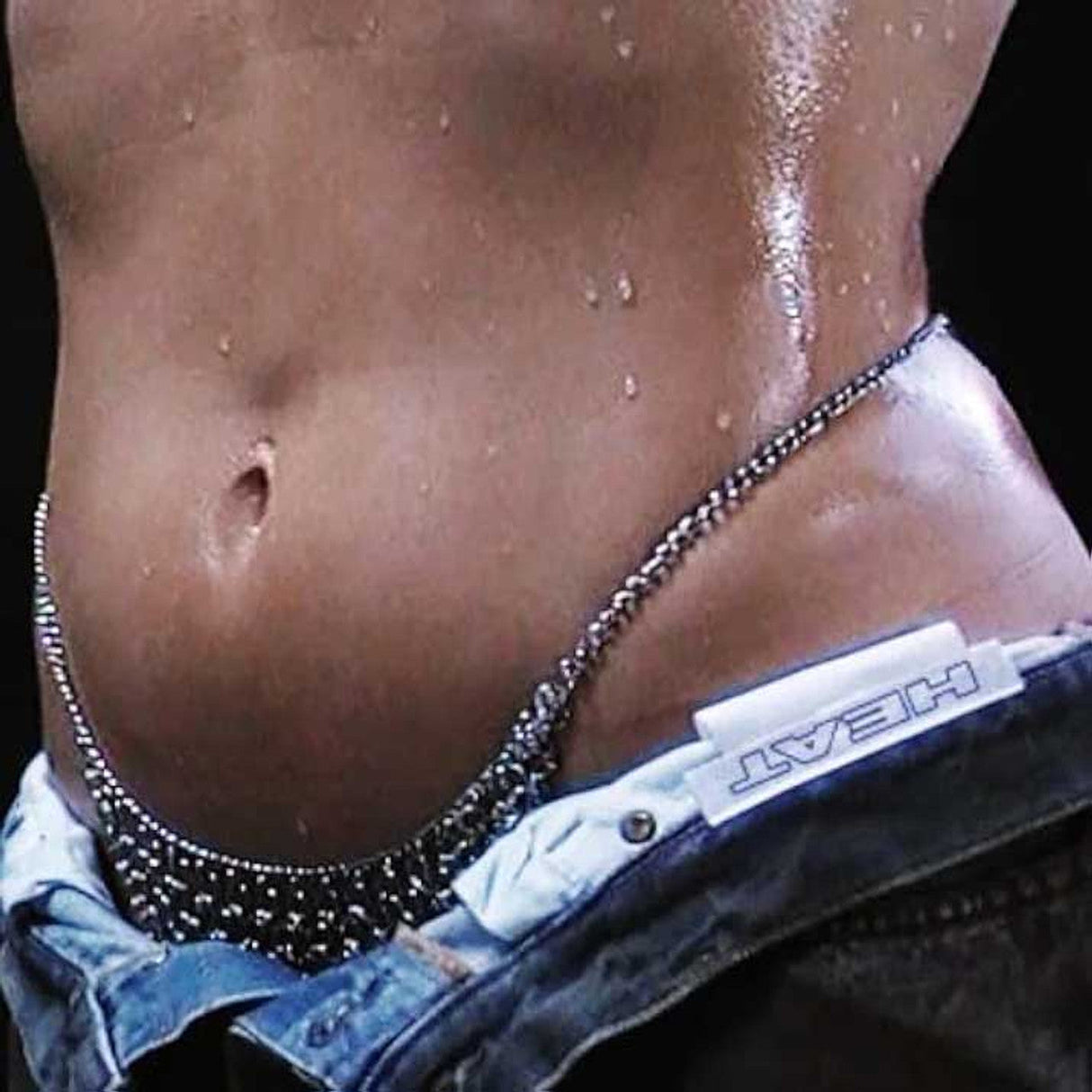Close-up of glistening torso with chain and HEAT label on denim