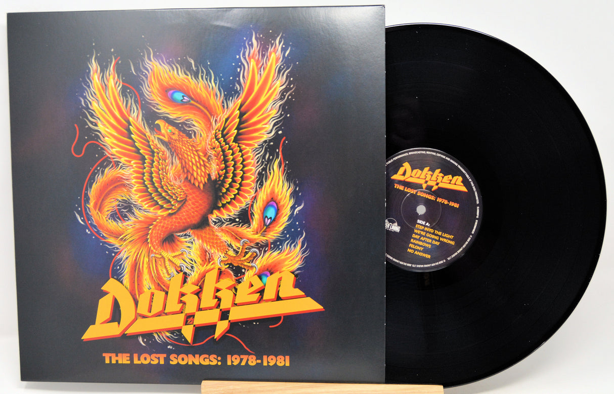A vinyl record and its album cover for Dokken's 'The Lost Songs: 1978-1981'. The cover features a vibrant orange and yellow phoenix illustration on a dark background, with the band's name in a bold yellow font.
